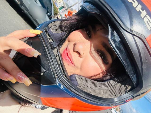 ChloeVixen wearing a black and orange motorcycle helmet, looking up with a slight smile, showing her face and yellow nail polish, sun shining on her.