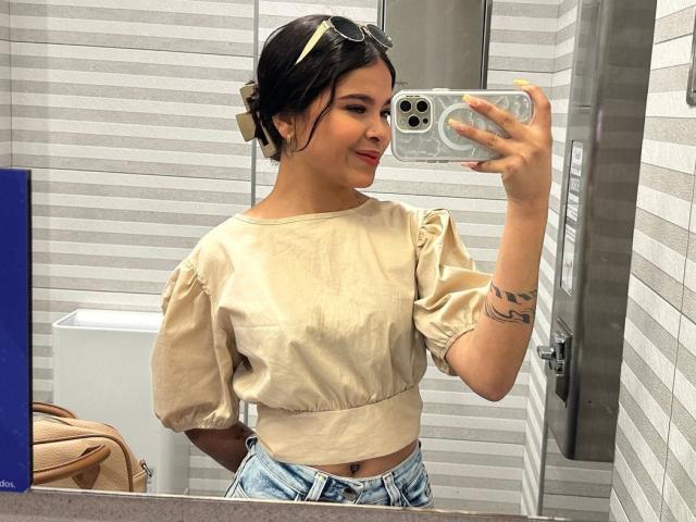 ChloeVixen with a cute smile, holding a phone in a bathroom mirror selfie. She's wearing a beige puffy shirt and blue jeans, with a tattoo on her left arm.