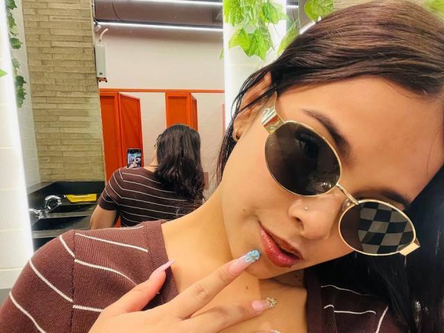 ChloeVixen with dark hair, wearing round sunglasses with a checker pattern, brown striped shirt, and blue glitter nail, touching her lips in a mirror selfie.