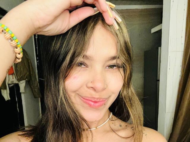 ChloeVixen with dirty brown hair, pink nose ring, subtle smile, and a pink lip mark, wearing a beaded bracelet and choker, touching her head with a manicured hand.
