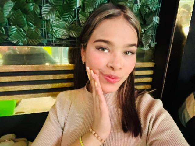 ChloeVixen has light skin, long brown hair, and is wearing a beige rib-knit sweater. She's pouting with her finger on her cheek, looking cute. Her nails are long.