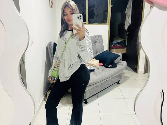 ChloeVixen with blonde hair, wearing a gray hoodie, black pants, and green shoulder bag, taking a mirror selfie, showing a cute ass, in a room with a gray couch.