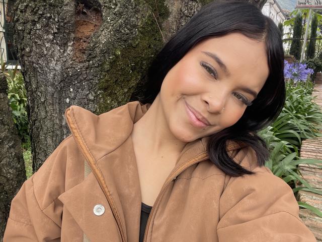 ChloeVixen with dark brown hair, light brown jacket, smiling slightly, small button on jacket, tree bark in background, green plant on right, casual selfie, cute, soft makeup.