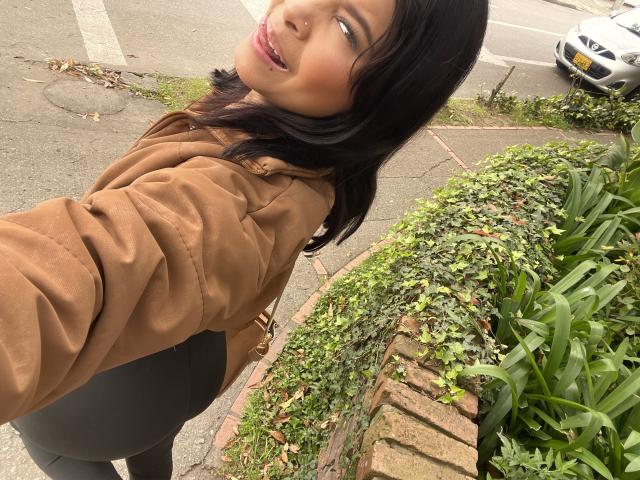 ChloeVixen with dark hair, brown coat, and black pants, looking back with a slight smile, her ass slightly raised, standing next to a brick wall with greenery.