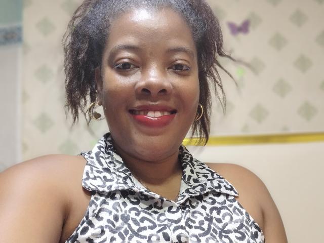 BrownieBabe with dark brown skin, curly black hair, red lipstick, hoop earrings, wearing a leopard print sleeveless top, smiling, showing her teeth. Her tits are partially visible.
