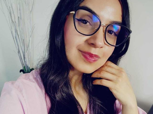 LizethWhite with long black hair, wearing pink shirt, black glasses with blue reflection, pink lipstick, smiling, hand near chin, looking at camera, slightly shiny skin.