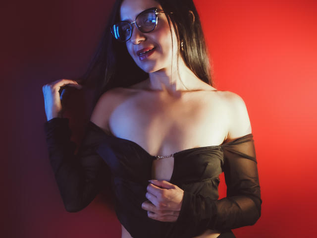 LizethWhite with glasses, black off-shoulder top, exposing her tits, holding her hair, smiling, red background, her hand on her chest, looking sexy, light skin, dark hair.