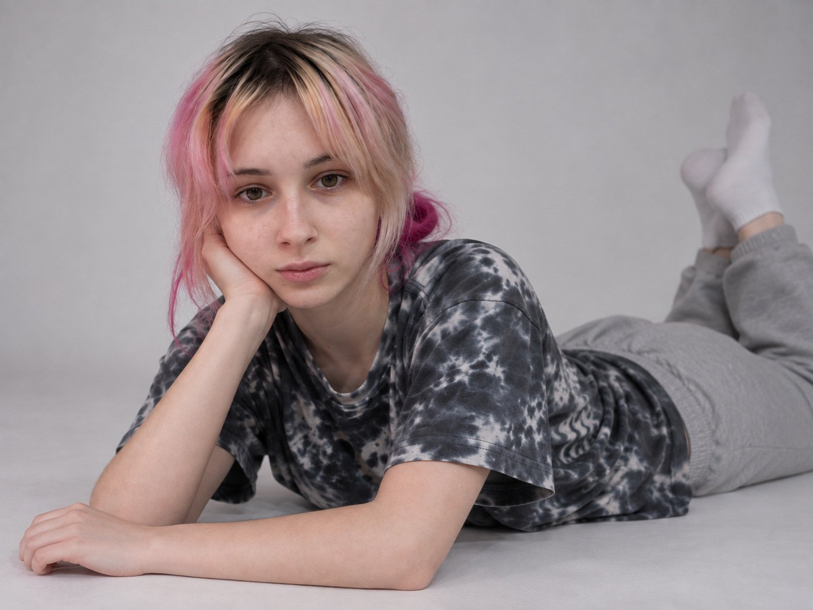 JulieYerry, com cabelo rosa e loiro, deitada de bruços, vestindo uma camiseta tie-dye e calças cinzas, olhando para a câmera, com a mão no rosto, usando meias brancas, com uma expressão de tédio..