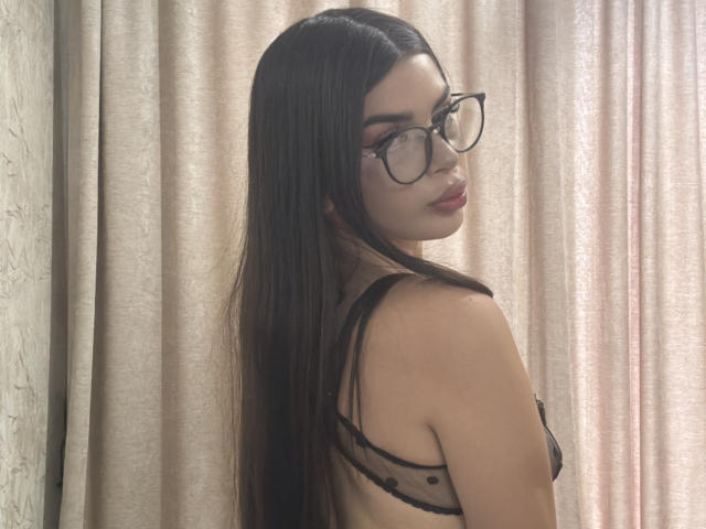 LilithMorrigan69, avec de longs cheveux noirs, des lunettes et du rouge à lèvres, portant un soutien-gorge noir transparent, montrant ses petites seins et son dos nu, regardant par-dessus son épaule..