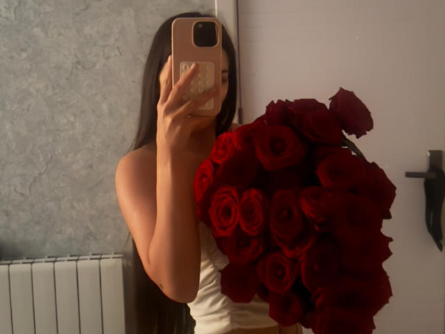 LilithMorrigan69 met lang donker haar, die een grote bos rode rozen vasthoudt, gekleed in een witte jurk zonder bandjes, die een spiegelzelfportret maakt met haar roze telefoon..