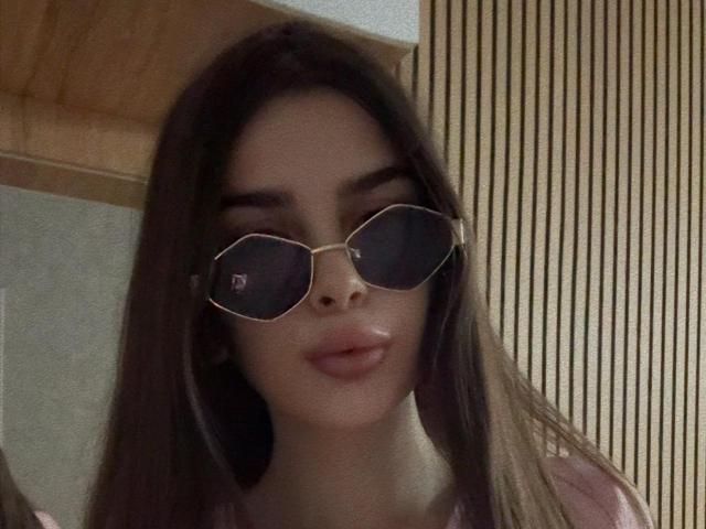 LilithMorrigan69 with long brown hair, wearing dark sunglasses, has full pink lips and a light skin tone. She's in a pink shirt, looking slightly to the side.