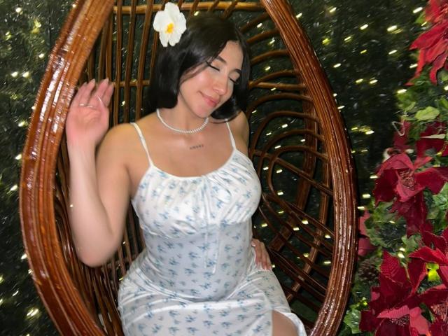 MaiteCollins, com cabelo preto, uma flor branca no cabelo, vestindo um vestido azul com estampas florais, sentada em uma cadeira de vime, com a mão levantada, sorrindo, mostrando os seios, usando um colar e com uma tatuagem..
