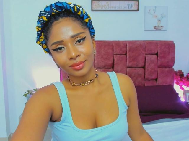 MeganWlliams with brown skin, blue and yellow headscarf, light blue tank top, chain necklace, and pink lipstick, showing her tits and a subtle smile in a bedroom.