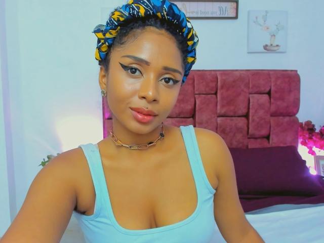 MeganWlliams with dark skin, blue headscarf, yellow and blue pattern, wearing a light blue tank top, showing her tits, wearing a gold chain, and eyeliner, smiling.