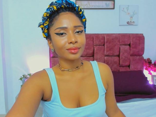 MeganWilliams has tan skin, curly blue and yellow hair, wearing a blue tank top, showing her tits, with winged eyeliner, a choker, and a subtle smile.