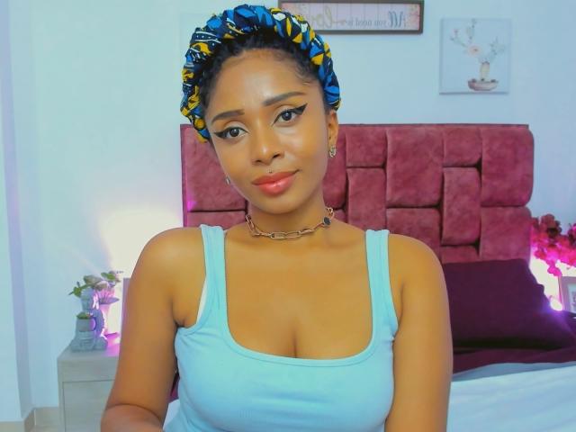 MeganWilliams with brown skin, blue patterned headwrap, blue tank top, showing her tits, wearing a chain necklace, smiling, in a bedroom with pink headboard.