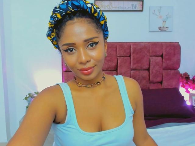 MeganWilliams with brown skin, blue eyes, and curly hair in a yellow and blue patterned headscarf, wearing a light blue tank top, showing her tits, smiling at the camera.