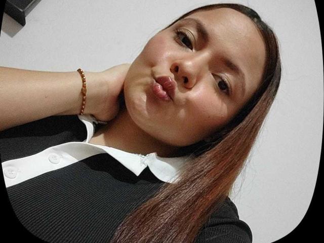 CherryScarlett has puffy lips, a brown bracelet, and long brown hair, wearing a black and white shirt, with a hand behind her head, looking slightly annoyed.