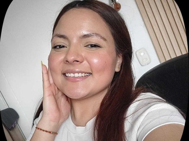 CherryScarlett ha una pelle luminosa, occhi marroni e un sorriso che mostra i denti. Sta toccando la guancia con una mano, indossa una camicia bianca e ha lunghi capelli castani..