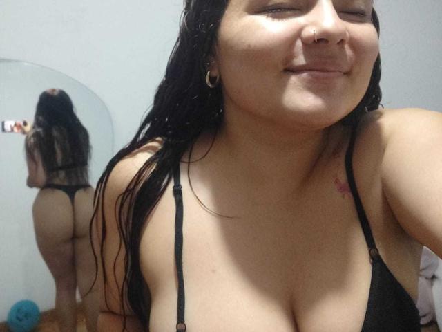CherryScarlett with wet black hair, smiling, wearing a black bra, showing her tits and small pink tattoo on her right shoulder. Her butt is visible in a mirror.