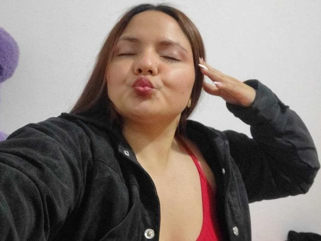 CherryScarlett, with brown hair, pouty lips, and eyes closed, blows a kiss while touching her ear. She wears a black jacket over a red top, showing her tits.