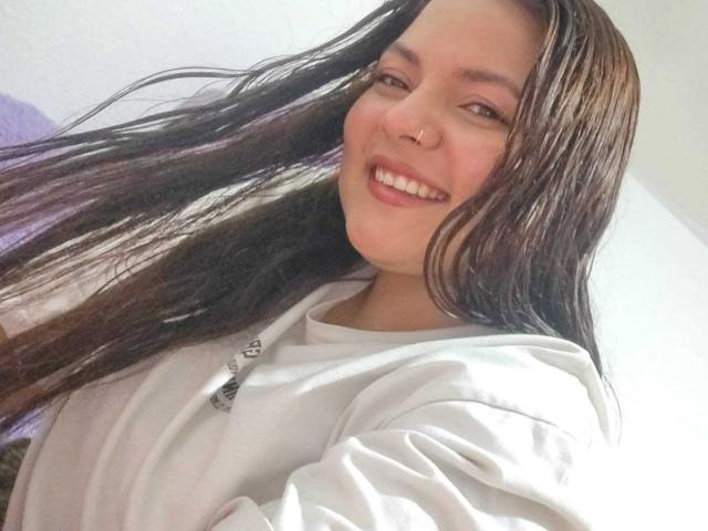CherryScarlett with long, wet brown hair, smiling, wearing a white hoodie, nose ring, and white teeth, looking at the camera, in a bright room.