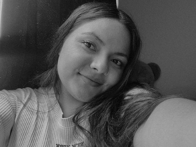 Black-and-white selfie of CherryScarlett with long, straight hair, smiling, nose ring, wearing a ribbed shirt, leaning on a surface, soft light, relaxed expression.