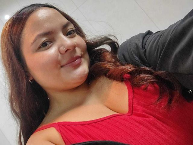 CherryScarlett, con i capelli castano ondulati, indossa una maglietta a coste rossa che mette in mostra il seno, sorride leggermente, e indossa una giacca nera, un anello al naso e piccoli orecchini a cerchio, in un selfie..