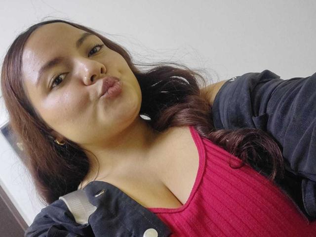 CherryScarlett with puffy lips, brown wavy hair, wearing a pink ribbed top and black jacket, showing her big tits and a strap from a bag.