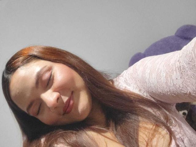 CherryScarlett with closed eyes, slight smile, and brown hair, wearing a pink lace top, sunlit face, and a purple stuffed animal in the background.