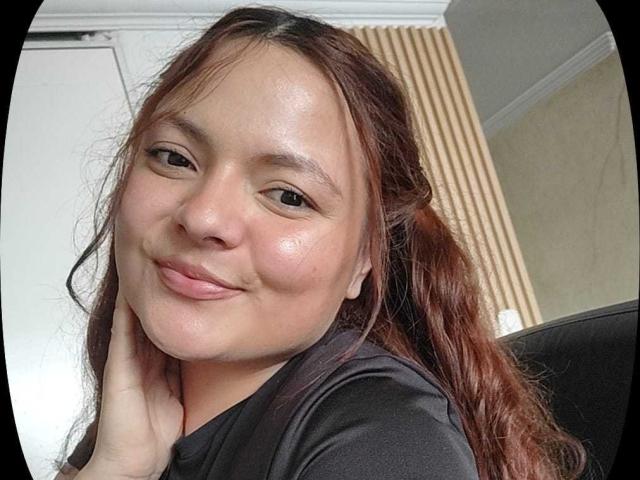 CherryScarlett with brown wavy hair, slightly shiny skin, and a subtle smile, wearing a black shirt, touching her neck, showing small, cute tits, and a relaxed, confident look.