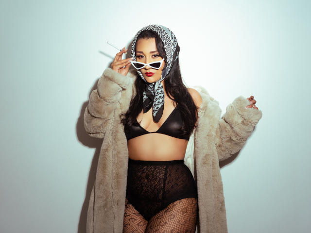Kourtney Parker con un sujetador negro, tanga de encaje de cintura alta, medias de rejilla, abrigo de piel beige, máscara blanca y bandana, sosteniendo un cigarrillo, mostrando sus pechos y glúteos..