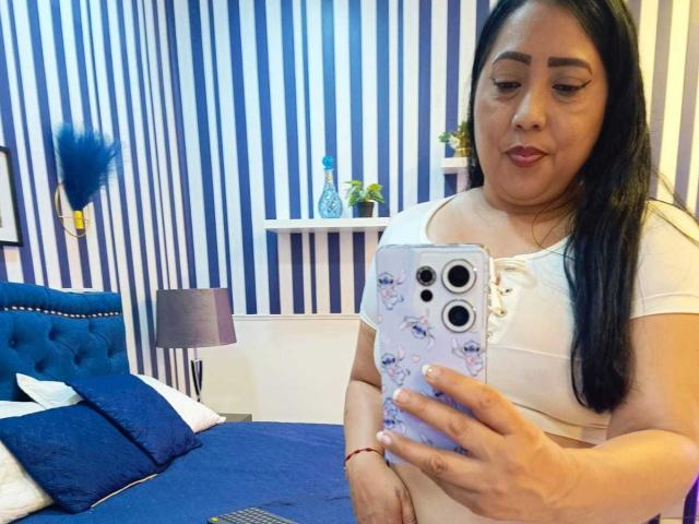 AmberMoretti, chubby with long black hair, wearing a white crop top, holding a floral phone case, taking a mirror selfie, showing her tits and pink lipstick.