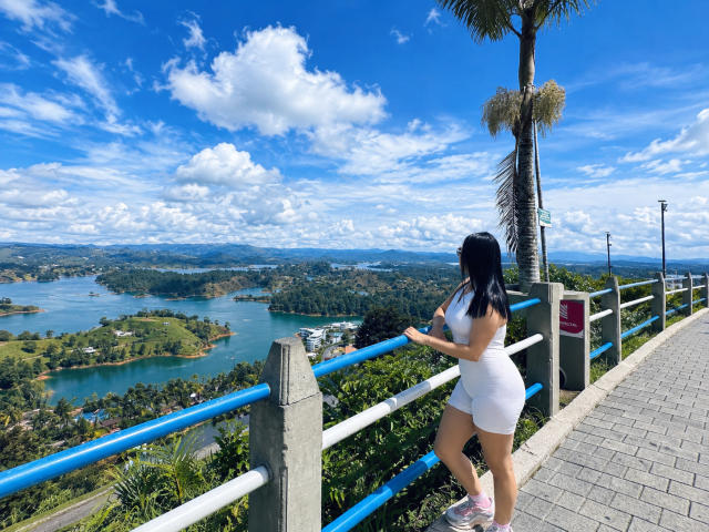 MiaAylen with black hair, white tight dress, and pink sneakers, standing on a blue and white railing, looking at a lush, blue-watered landscape under a bright, cloudy sky.