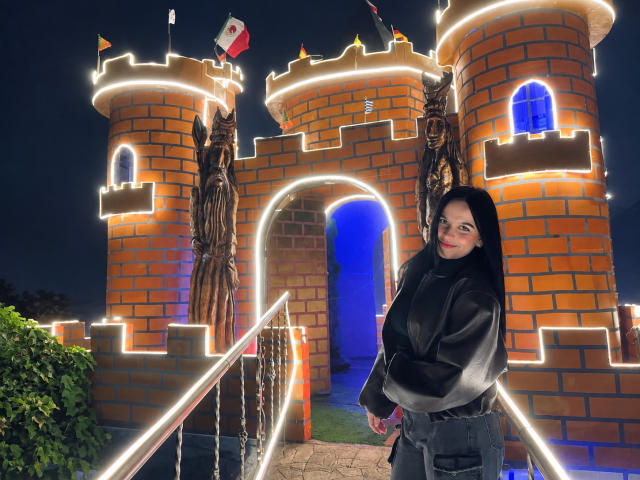 MiaAylen stands in front of a brick castle illuminated by white lights, wearing a black shiny coat and long black hair, with a confident smile. Night scene.