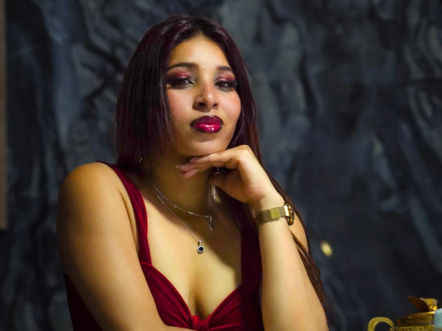 JaneKetty with dark red hair, pink lipstick, and red dress, touching her chin, wearing a gold bracelet, necklace with a small pendant, and dark makeup.