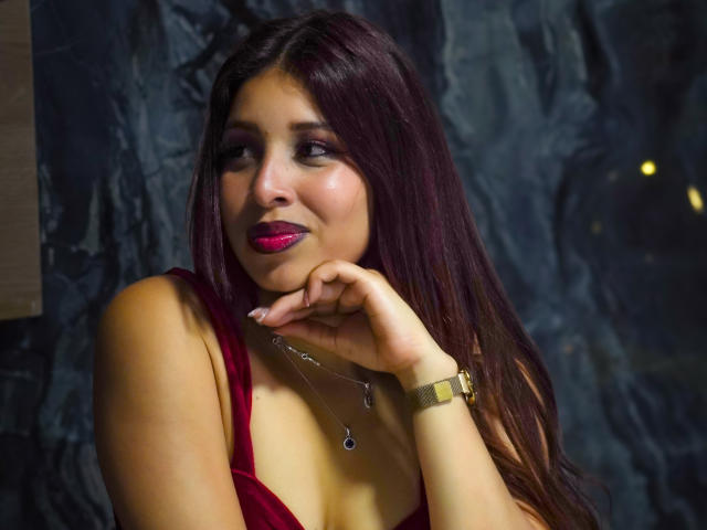 JaneKetty with dark red hair, pink lipstick, and a gold watch, resting her chin on her hand, wearing a red dress, and a silver necklace with a small pendant.