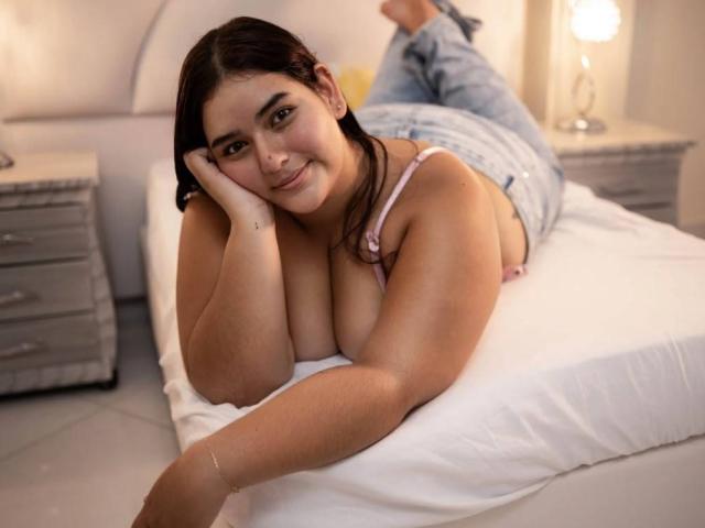 MiaWialliams, sdraiata su un letto, con il suo seno prosperoso e il suo sorriso dolce, indossa un reggiseno rosa e jeans blu, con la testa appoggiata alla mano e le gambe sollevate..