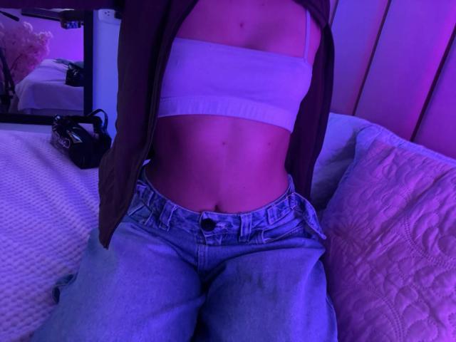 HannaThorne with a flat white sports bra, showing her tits, and wearing blue jeans, her brown jacket open, in purple-lit bedroom, sitting on a bed.