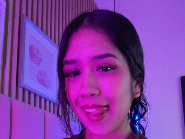 HannaThorne with dark hair, pink and purple lighting, glossy lips, and blue ear cuff, smiling with tongue slightly out, looking directly at camera.