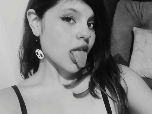 SophieHill with dark hair, nose ring, and skull earrings, sticking her tongue out, showing off her eyeliner, and wearing a black spaghetti strap top. Black and white selfie.