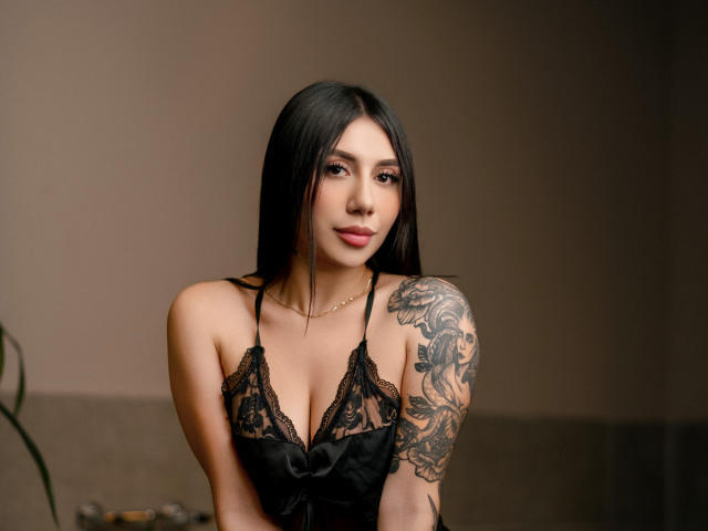 AllanaMoon with long black hair, lace black dress, showing tattoos on her right arm, and a subtle gold necklace, looking at the camera with a slight smile.