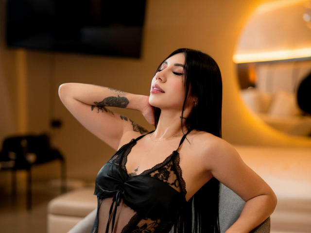 AllanaMoon with long black hair, eyes closed, wearing black lace lingerie, has her arm behind her head, showing a tattoo on her arm, and a relaxed, sexy expression.