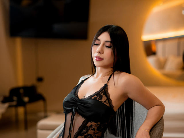 AllanaMoon with long black hair, wearing a black lace lingerie top, showing her small tits, sitting in a chair, eyes closed, in a softly lit room.