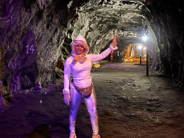 Mara Roberts, portant un sweat à capuche blanc, une écharpe rose, un pantalon argenté brillant et des bottes blanches, se tient dans une grotte sombre, lève un bras et sourit. Ses seins sont petits..