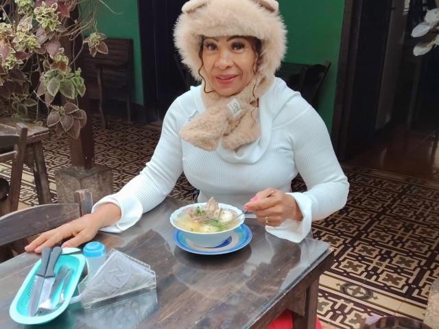 MaraRoberts with fluffy beige hat, white sweater, eating soup from a blue bowl, holding spoon, wooden table, patterned floor, plant in background, smiling, hands on table, napkin.