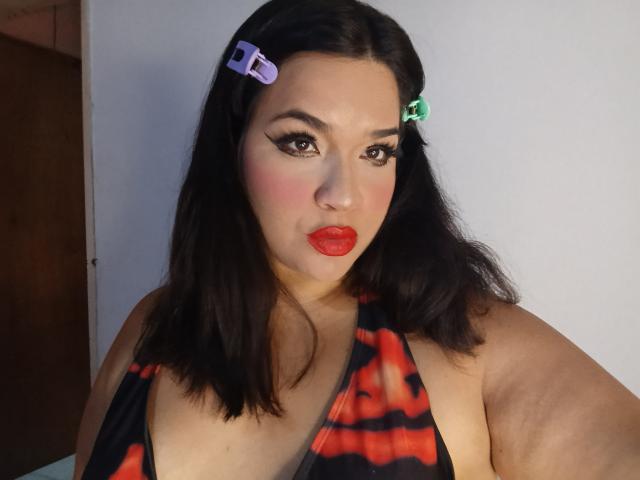 AntonelaPark with dark hair, red lipstick, and black-red patterned top, wearing purple and green hair clips, has bold eyeliner and blushed cheeks, showing her tits slightly.