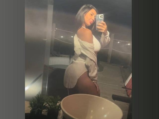 NahomyStone in a loose white shirt, showing her big ass and thick thighs in a mirror selfie, holding a phone with a blue case, in a dimly lit room.