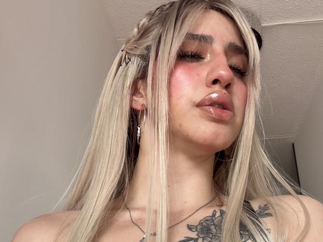 ElodieRoussee with blonde hair, braided side, glossy pink lips, red blush on cheeks, visible tattoos on chest, long eyelashes, wearing black cat ears, looking up, slightly parted lips.