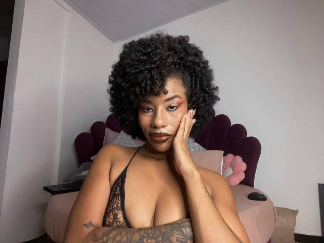 KarlaRicochet, con cabello negro rizado, piel morena y tatuajes, vestida con una blusa negra de encaje, apoyando su barbilla en su mano, mostrando sus pechos. Está mirando a la cámara..
